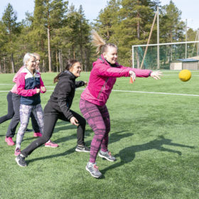 Neljä nuorta naista seisoo jonossa. Ensimmäinen heittää keltaista palloa. Muut kurkistavat edellä olevien selkien takaa.