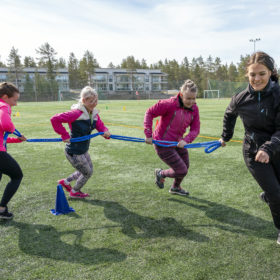 Neljä naista juoksee niin, että heidät on kiinnitetty peräkkäin sinisellä köydellä. Naiset kiertävät sinistä maassa olevaa tötteröä.