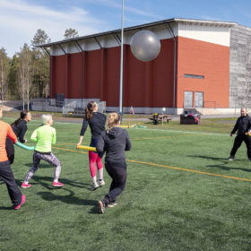 Joukko nuoria naisia pelaa jalkapalloa isolla harmaalla jumppapallolla. Naiset juoksevat pitäen käsissään vaahtomuovista pötkylää.