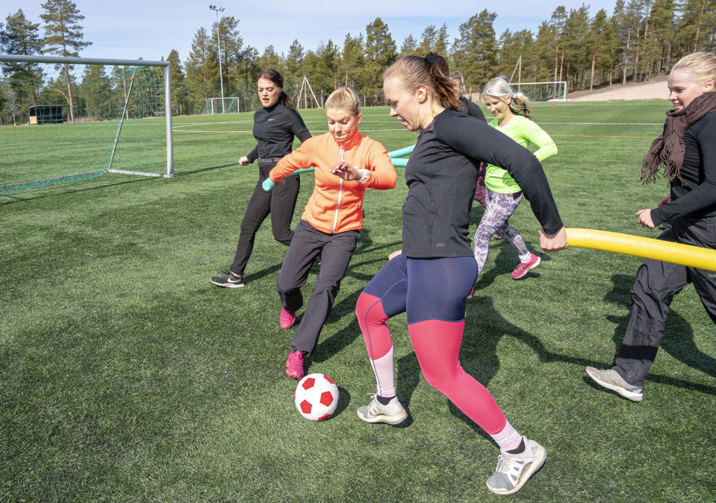 Joukko nuoria naisia pelaa jalkapalloa pehmeällä puna-valkoisella jalkapallolla. Naiset juoksevat pitäen käsissään vaahtomuovista pötkylää.