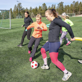 Joukko nuoria naisia pelaa jalkapalloa pehmeällä puna-valkoisella jalkapallolla. Naiset juoksevat pitäen käsissään vaahtomuovista pötkylää.