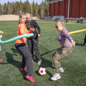 Joukko nuoria naisia pelaa jalkapalloa pehmeällä puna-valkoisella jalkapallolla. Naiset juoksevat pitäen käsissään vaahtomuovista pötkylää.