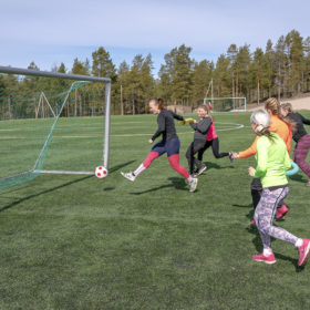 Joukko nuoria naisia pelaa jalkapalloa pehmeällä puna-valkoisella jalkapallolla. Naiset juoksevat pitäen käsissään vaahtomuovista pötkylää.