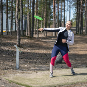 Urheilullisiin vaatteisiin pukeutunut nainen heittää vihreää frisbeekiekkoa. Takana näkyy havupuiden runkoja.