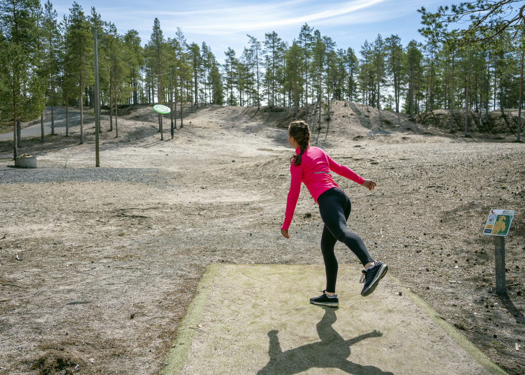 Tummahiuksinen, pinkkitakkinen nainen heittää frisbeetä kohti kauempana häämöttäviä havupuita.