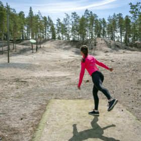 Tummahiuksinen, pinkkitakkinen nainen heittää frisbeetä kohti kauempana häämöttäviä havupuita.