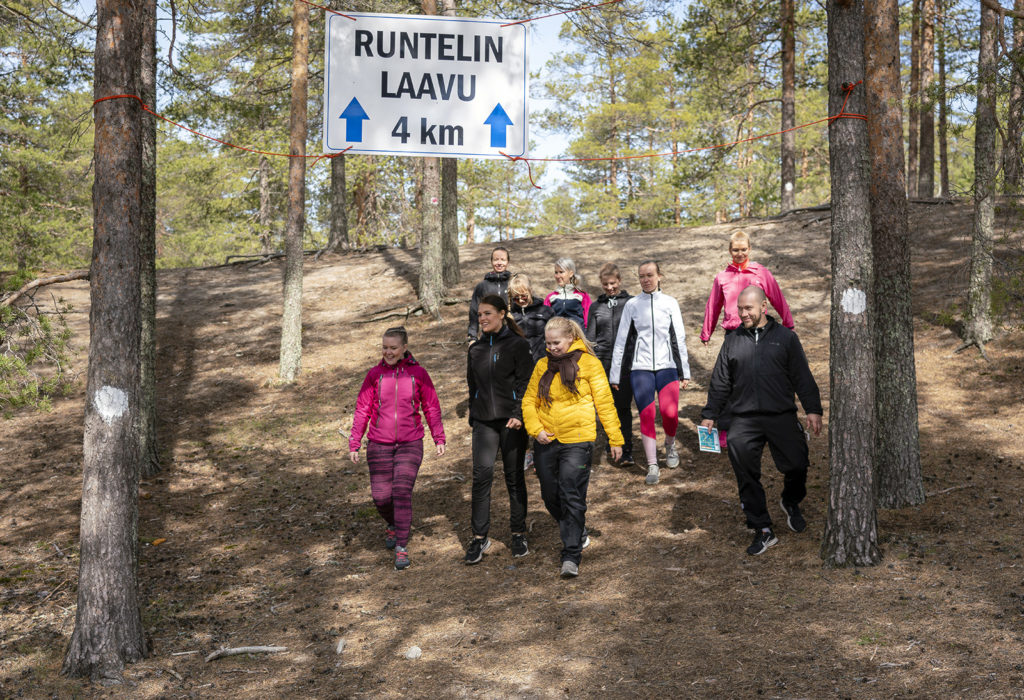 Joukko ulkoiluvaatteisiin pukeutuneita henkilöitä laskeutuu alas jyrkkää rinnettä. Heidän yläpuolellaan on kultti, jossa lukee Runtelin laavu 4km. Kyltissä on lisäksi kaksi nuolta ylöspäin.