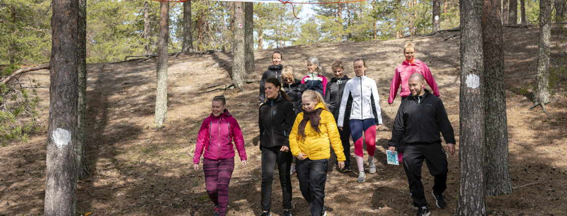 Joukko ulkoiluvaatteisiin pukeutuneita henkilöitä laskeutuu alas jyrkkää rinnettä. Heidän yläpuolellaan on kultti, jossa lukee Runtelin laavu 4km. Kyltissä on lisäksi kaksi nuolta ylöspäin.