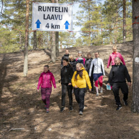 Joukko ulkoiluvaatteisiin pukeutuneita henkilöitä laskeutuu alas jyrkkää rinnettä. Heidän yläpuolellaan on kultti, jossa lukee Runtelin laavu 4km. Kyltissä on lisäksi kaksi nuolta ylöspäin.