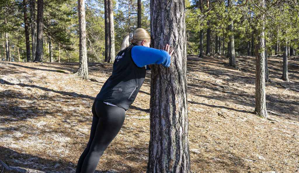 Vaaleahiuksinen nainen nojaa käsillään puunrunkoon. Taustalla hiekkapohjainen metsä, jossa isoja puita.