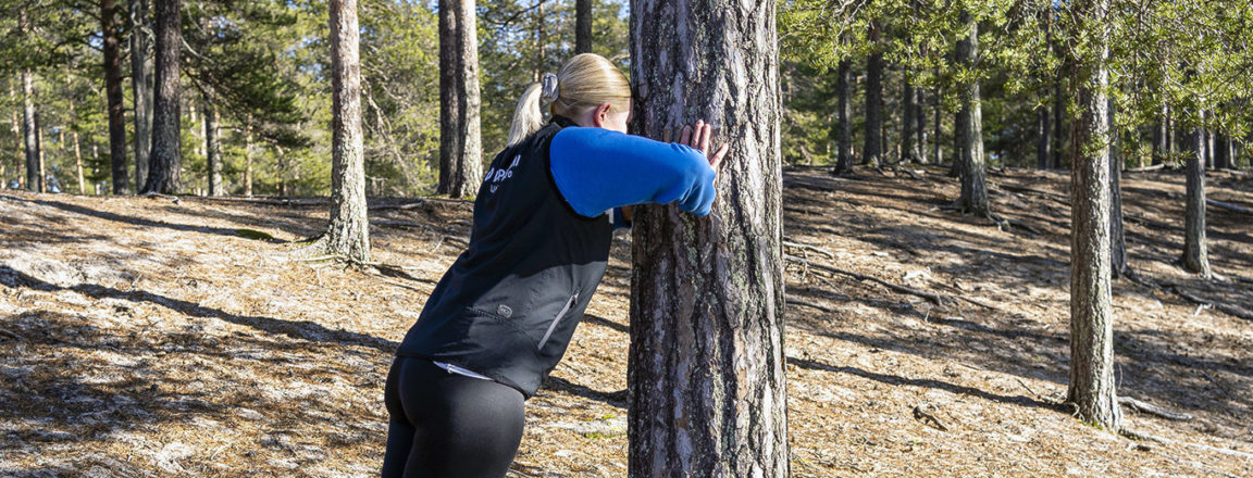 Vaaleahiuksinen nainen nojaa käsillään puunrunkoon. Taustalla hiekkapohjainen metsä, jossa isoja puita.