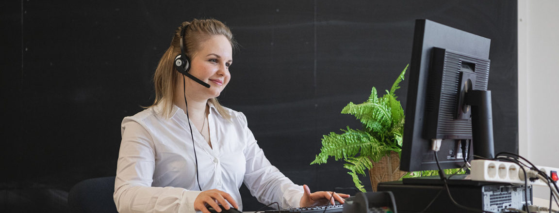 Vaaleahiuksinen nuori nainen istuu tietokoneen ääressä. Naisella on kuuloke-mikrofoni headset päässään. Pöydällä on tietokoneen rinnalla kassapääte ja viherkasvi.