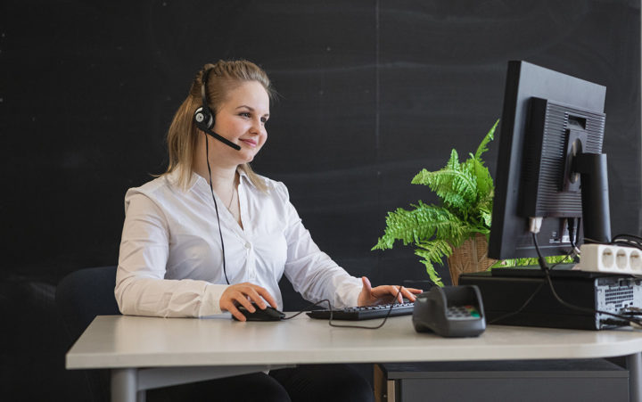 Vaaleahiuksinen nuori nainen istuu tietokoneen ääressä. Naisella on kuuloke-mikrofoni headset päässään. Pöydällä on tietokoneen rinnalla kassapääte ja viherkasvi.