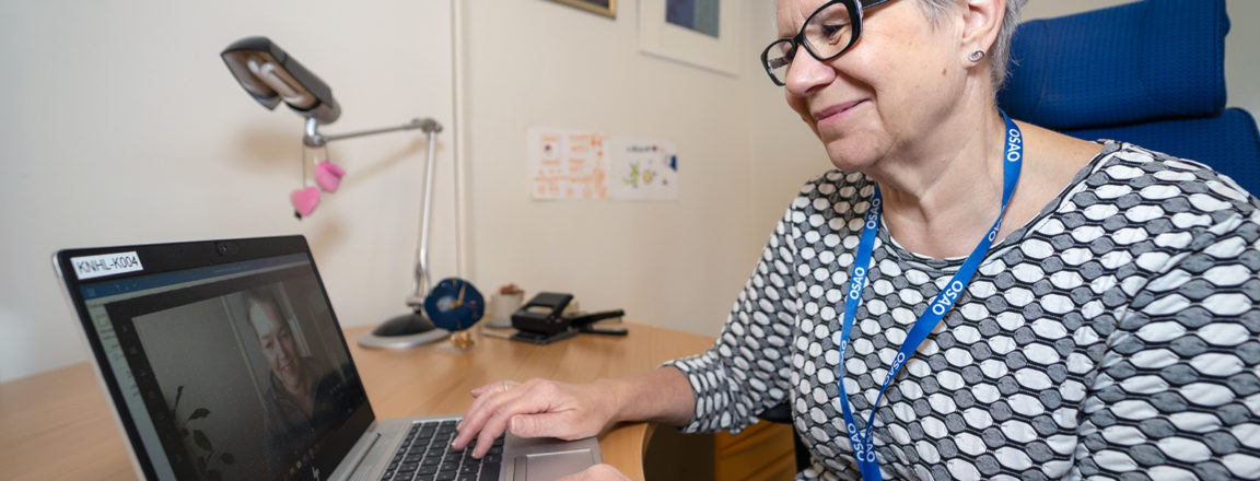 Musta-valkoiseen paitaan pukeutunut lyhythiuksinen ja silmälasipäinen nainen istuu työpöydän äärellä ja katsoo hymyillen tietokonetta. Tietokoneen ruudulla näkyy kuva naishahmosta, jonka kanssa tietokoneella työskentelevä nainen on yhteydessä etäyhteydellä.