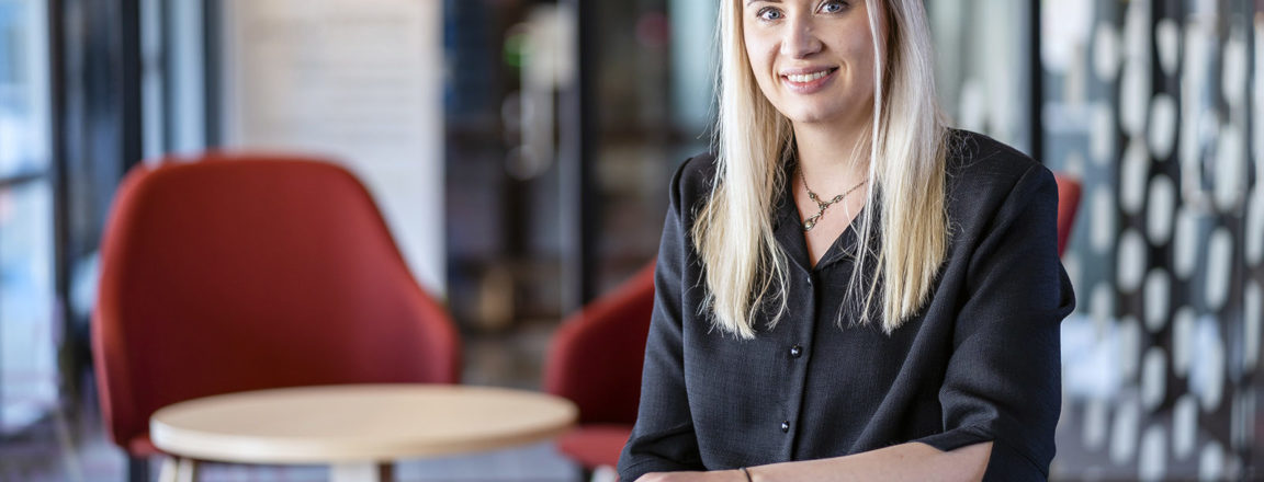 Vaalea, pitkähiuksinen nainen istuu pöydän ääressä. Naisella on tummanharmaa paita päällä. Takana näkyy punainen nojatuoli ja pieni pyöreä pöytä.