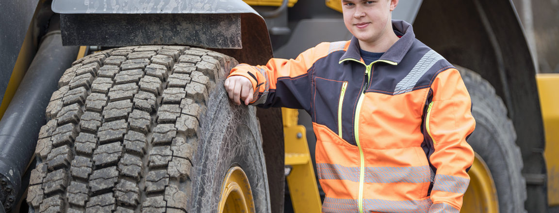 Oranssiin työtakkiin pukeutunut lippalakkipäinen nuori mies nojaa suuren pyörökoneen renkaaseen.