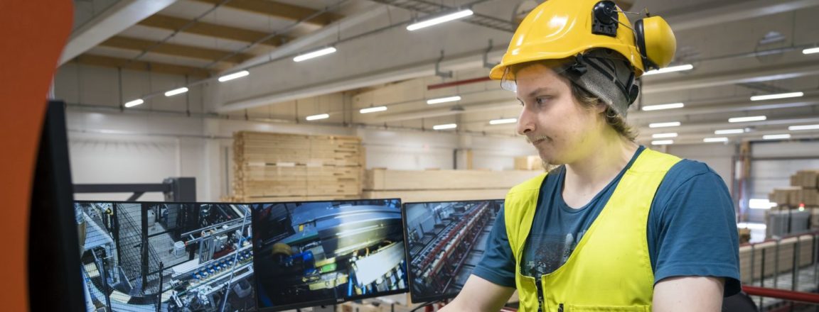 Keltaiseen liiviin ja suojakypärään pukeutunut mies seisoo korokkeella, jossa on oranssi kaide. Mies tarkkailee isoja monitoreja, jotka ovat pöydällä. Monitoreissa näkyy teollisuuden linjaston eri vaiheita.