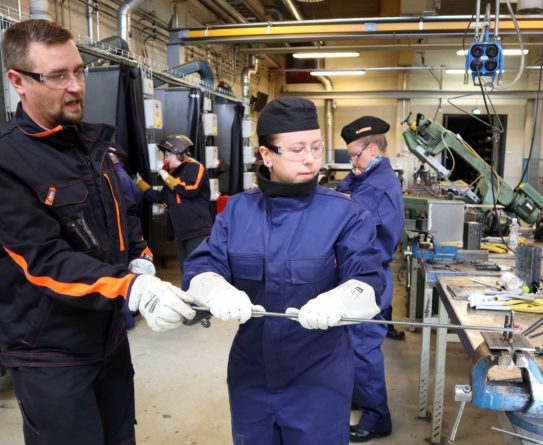 Miesopettaja ohjaa työsalissa naisopiskelijaa metallin taivuttamisessa. Molemmat ovat pukeutuneet työvaatteisiin. Taustalla näkyy työsalin koneita ja laitteita sekä toinen opiskelija ja opettaja hitsausmaskissa.