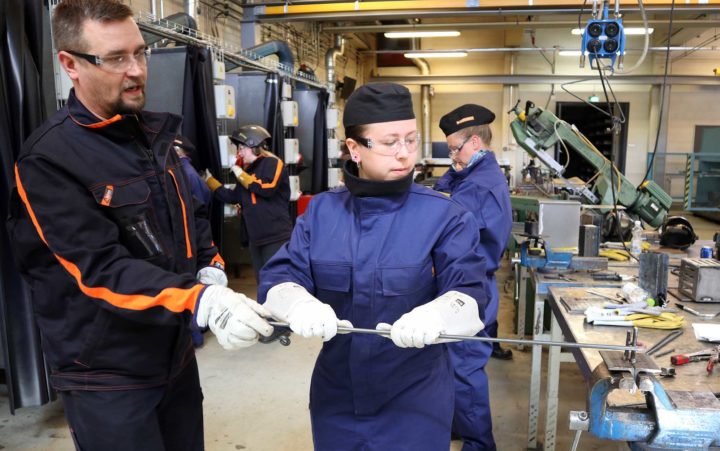 Miesopettaja ohjaa työsalissa naisopiskelijaa metallin taivuttamisessa. Molemmat ovat pukeutuneet työvaatteisiin. Taustalla näkyy työsalin koneita ja laitteita sekä toinen opiskelija ja opettaja hitsausmaskissa.