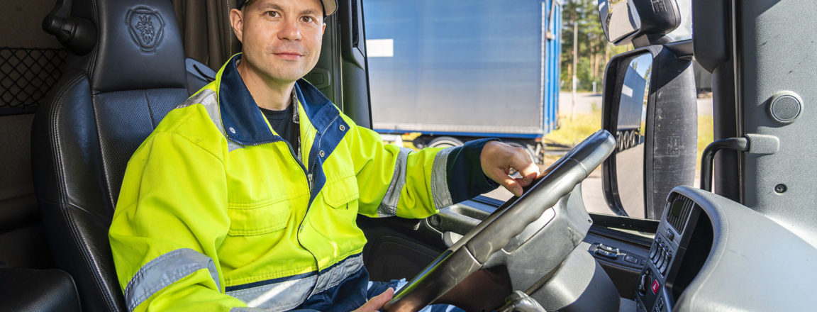 Keltaiseen huomiotakkiin pukeutunut mies istuu rekan ohjaamossa. Miehellä on päässään lippalakki, jonka lipan päälle on nostettu aurinkolasit. Mies istuu kuljettajan paikalla, jonka mustassa nahkapenkin nistatuessa on Scanian logo. Auton ikkunasta näkyy toisen rekan perävaunu, jossa on sinisen värinen liukuvärjäys.