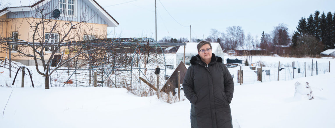 Tummaan, pitkään talvitakkiin pukeutunut nainen seisoo lumisessa pihassa. Naisen takana näkyy lehdettömiä pensaita ja harjakattoinen talo. Naisen vieressä on vaalea pystykorvainen koira.