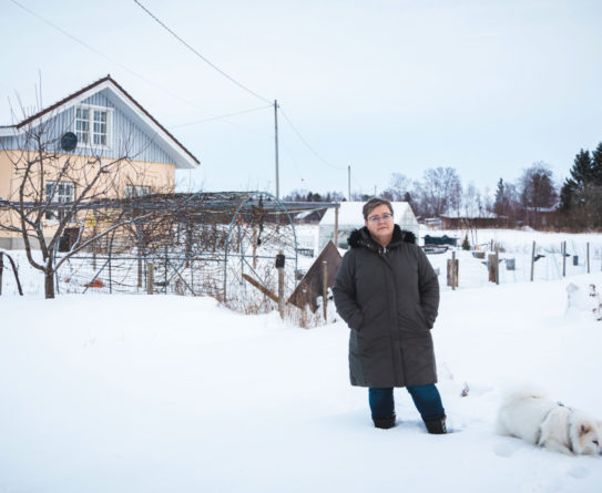 Tummaan, pitkään talvitakkiin pukeutunut nainen seisoo lumisessa pihassa. Naisen takana näkyy lehdettömiä pensaita ja harjakattoinen talo. Naisen vieressä on vaalea pystykorvainen koira.