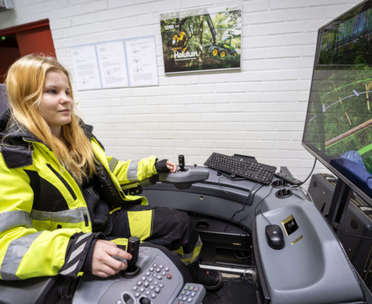 Keltaiseen huomioväriseen työtakkiin pukeutunut vaaleahiuksinen nuori nainen ohjaa metsäkonesimulaattoria.