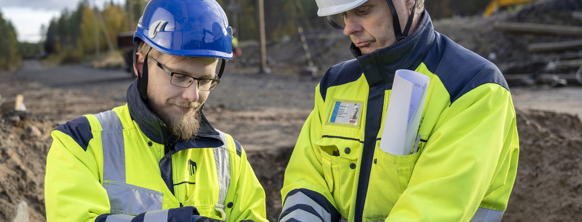 Kaksi keltaiseen työtakkiin pukeutunutta miestä seisoo kuopassa ja pitää käsissään sähköjohtoa. Oikeanpuoleisella miehellä on sininen kypärä ja hän kuorii johtoa pihdeillä. Oikeanpuoleisella miehellä on valkoinen kypärä.