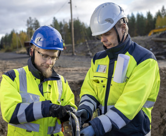 Kaksi keltaiseen työtakkiin pukeutunutta miestä seisoo kuopassa ja pitää käsissään sähköjohtoa. Oikeanpuoleisella miehellä on sininen kypärä ja hän kuorii johtoa pihdeillä. Oikeanpuoleisella miehellä on valkoinen kypärä.