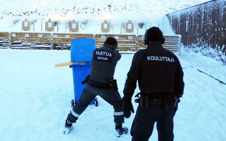 Harmaisiin haalareihin pukeutuneet henkilöt seisovat hajareisin ampumaradalla selin kameraan. Toisen henkilön selässä lukee Vartija, toisen selässä Kouluttaja.