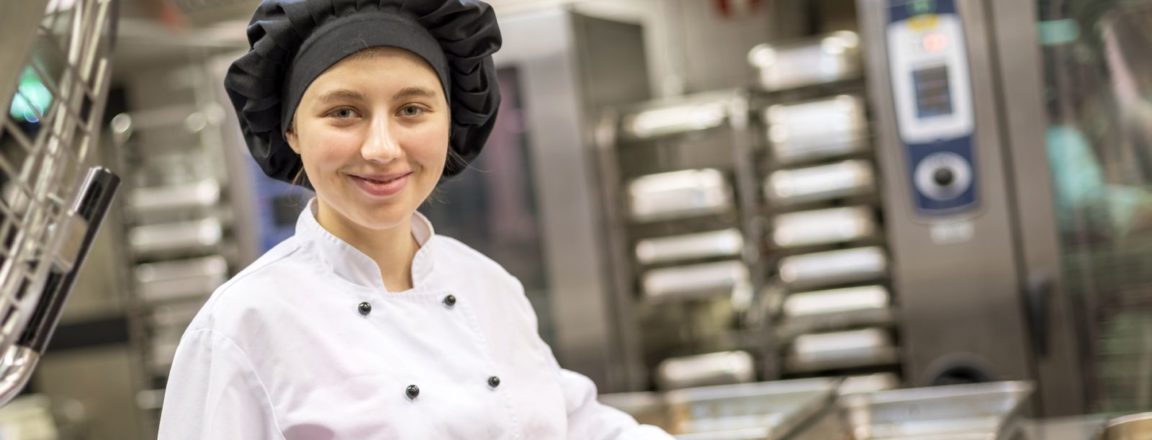 Valkoiseen työtakkiin ja mustaan hattuun pukeutunut nuori nainen seisoo ravintolan keittiössä ja nostaa kauhalla makaronipohjaista ruokaa astiasta.