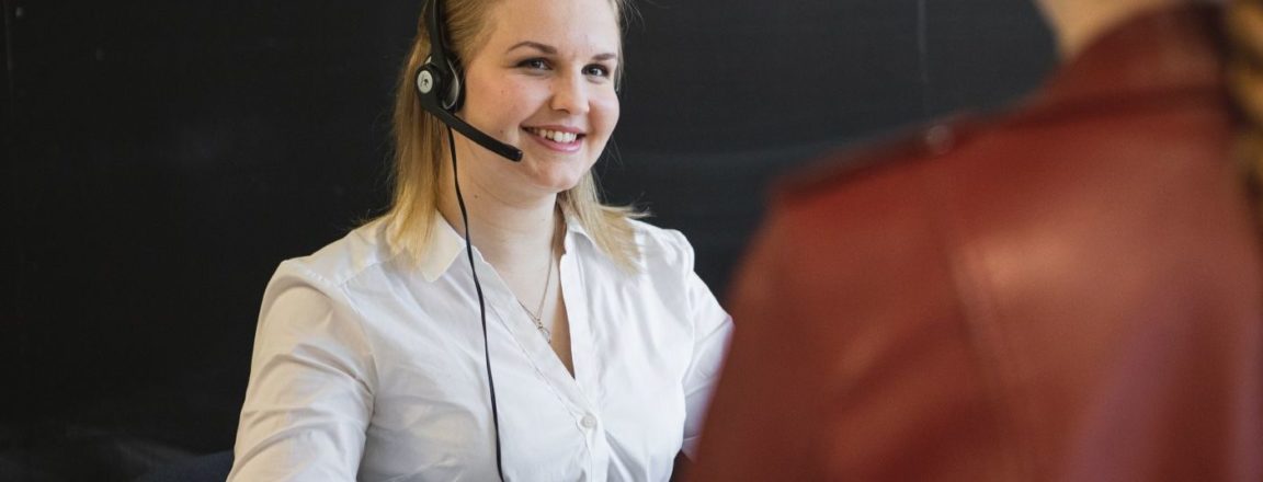 Vaaleahiuksinen, vaaleaan paitapuseoon pukeutunut nuori nainen on tietokoneella pöydän ääressä. Naisella on päässään mikrofonilla varustetut kuulokkeet. Naisen edessä on punaiseen takkiin pukeutunut asiakas.