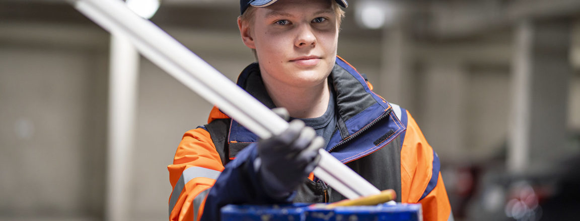 Sini-oranssiin takkiin ja siniseen lippalakkiin pukeutunut nuori mies seisoo tikkaiden takana ja pitää käsissään loisteputkea.