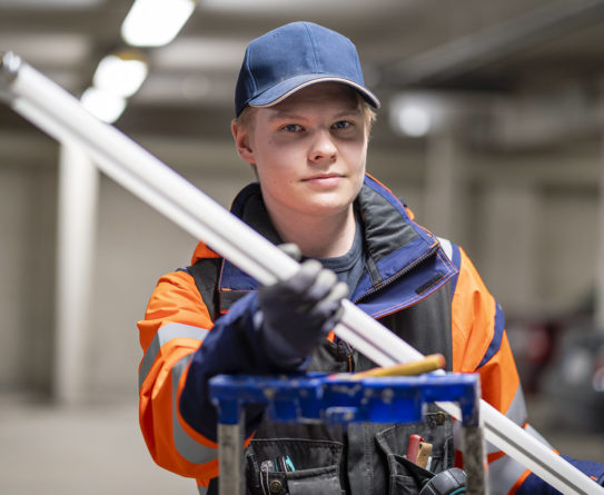 Sini-oranssiin takkiin ja siniseen lippalakkiin pukeutunut nuori mies seisoo tikkaiden takana ja pitää käsissään loisteputkea.