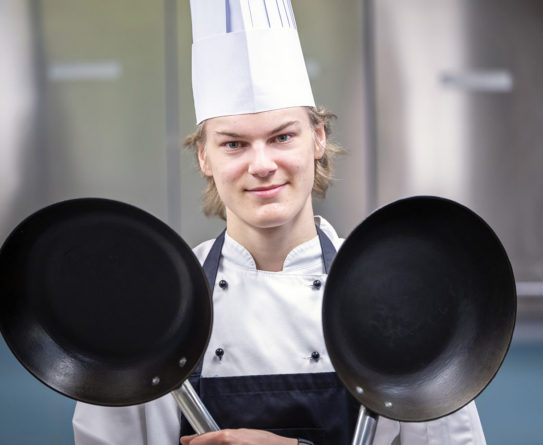 Valkoiseen kokinhattuun ja -takkiin pukeutunut nuori mies pitää molemmissa käsissään mustaa paistinpannua, jotka on nostettu pystyyn olkapäiden korkeudelle.