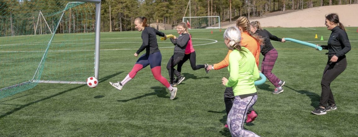 Joukko naisia pelaa nurmikentällä jalkapalloa.