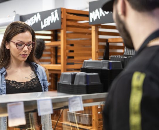 Nainen valitsee myymälän lämpötiskistä ruokaa. Häntä palvelee myymälän työntekijä, tummasilmäinen ja partainen mies.