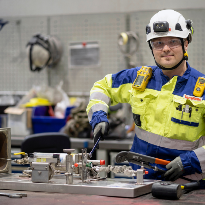 Mies huomiovärisissä työvaatteissa ja valkoisessa turvakypärässä katsoo kohti kameraa ja pitää isoa jakoavainta kädessä.