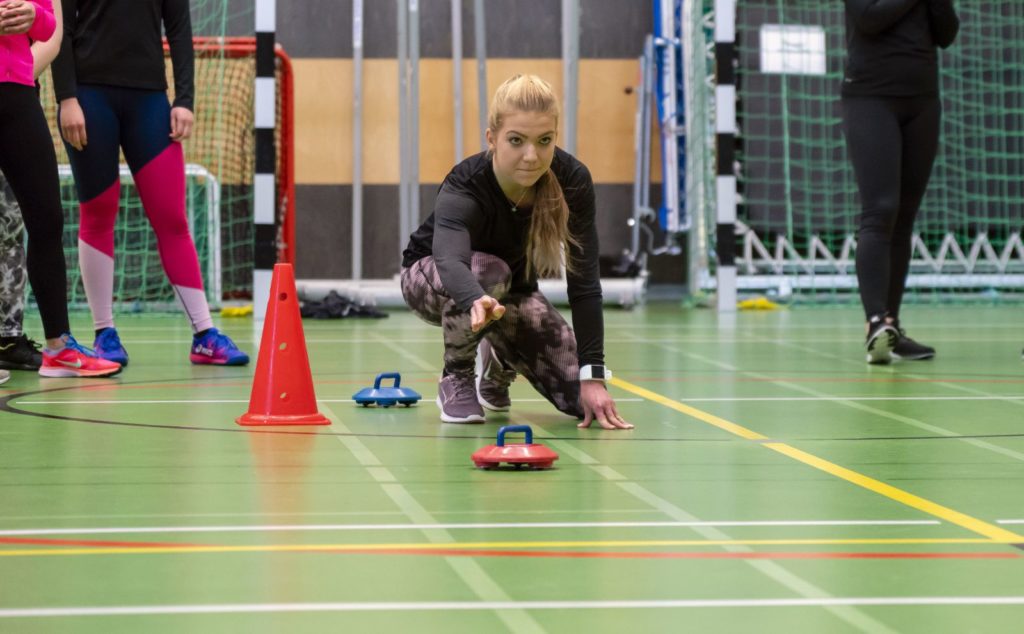 Vaaleahiuksinen nuori nainen työntää liikuntasalissa sisäcurlingin kiekkoa.