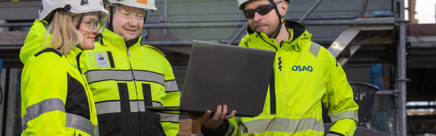 Kolme työvaatteisiin ja kypäröihin pukeutunutta henkilöä seisoo vierekkäin ja katsoo hymyillen yhden henkilön esittelemää kuvaa kannettavalta tietokoneelta. Taustalla seinä, jossa rakennustelineet.