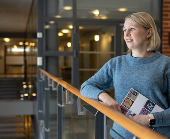 Utuisen siniseen neulepaitaan pukeutunut vaaleahiuksinen nainen nojaa kaiteeseen kirja kädessään.