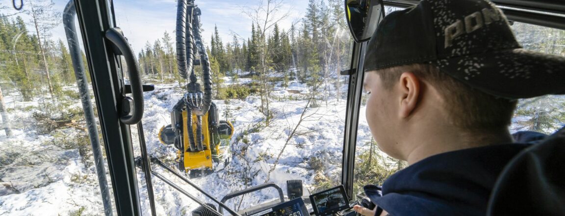 Lippalakkipäinen mies istuu metsäkoneen hytissä ja nostaa puita metsäkoneen kauhalla.