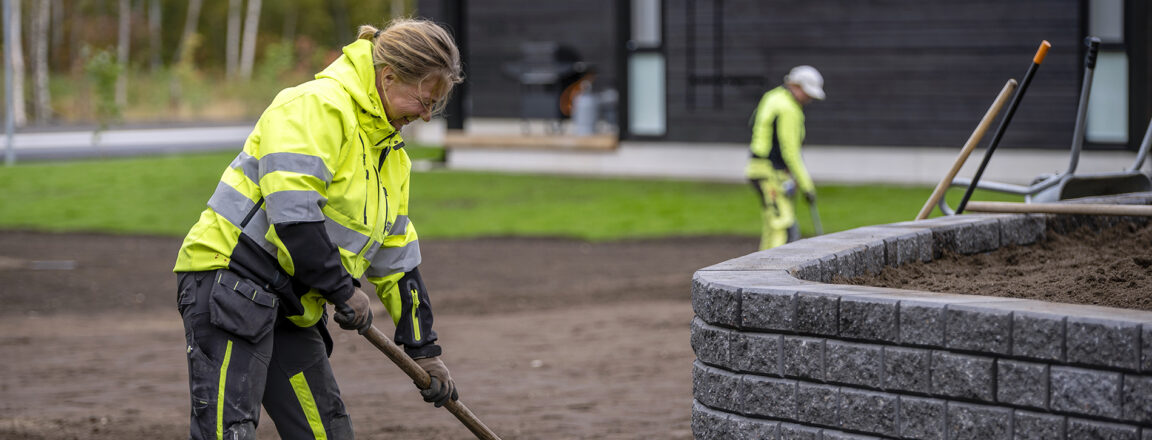 Nainen keltaisissa työvaatteissa levittää multaa juuri rakennetulle virheralueelle. Edessä kivimuuri.