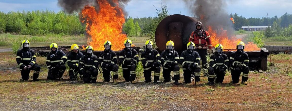 Joukko sammutustehtäviin soveltuviin asuihin pukeutuneita henkilöitä on polviseisonnassa kahden suuren nuotion edessä.