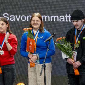 Kolme nuorta poseeraa iloisina palkintojen jaossa mitalit kaulassa ja kukkakimput kädessä.