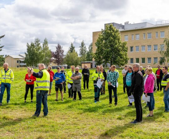 Ihmisiä seisoo nurmialueella, iso tiilirakennus taustalla.