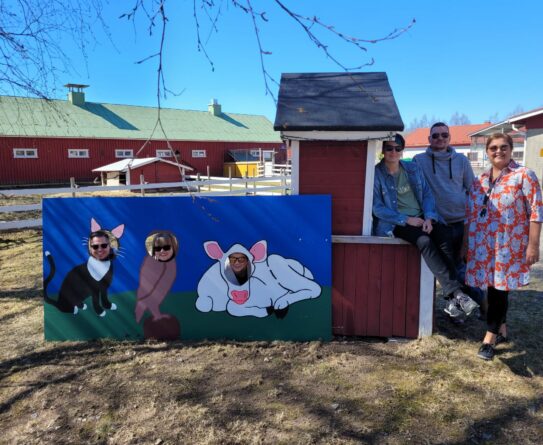 Kolme henkilöä kurkistaa tintamareskin pääaukoista, joista yksi on kissa, toinen lintu ja kolmas lehmä. Vieressä kolme muuta henkilöä poseeraa kameralle nojaten punaiseen maitolaituriin. Taustalla näkyy navetan piha-aluetta.