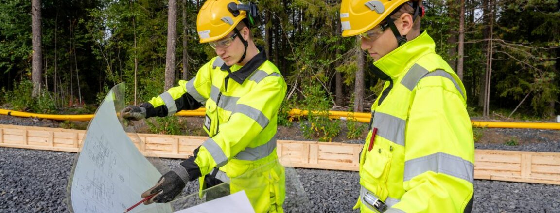 Kaksi talonrakentajaa tarkastelevat rakennuspiirustuksia. henkilöillä on työasut päällä ja takana kesäinen mäntymetsä.