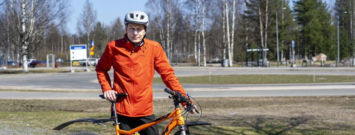 Pyöräilykypärään ja ulkoiluvaatteisiin pukeutunut Antti Rovamo seisoo oranssin maastopyörän kanssa pyörätiellä.