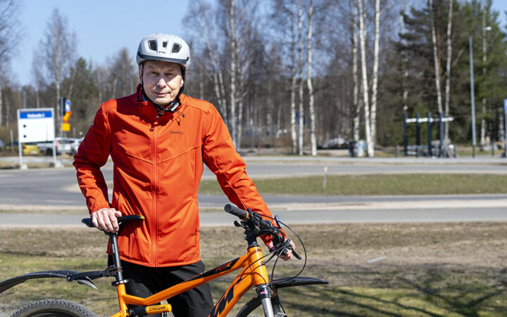 Pyöräilykypärään ja ulkoiluvaatteisiin pukeutunut Antti Rovamo seisoo oranssin maastopyörän kanssa pyörätiellä.